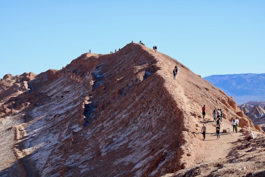 A rocky ridge resembling a serrated edge with several people walking and standing along the narrow path. The land formations have a reddish-brown hue, contrasting against the bright blue sky. The landscape appears arid and desert-like, set against distant mountain ranges.