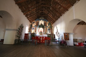 A wooden-roofed interior with an altar adorned with red and gold fabric. There are religious statues and symbols, with greenery such as palm branches adding to the decoration. The setting appears to be a simple but reverent space, with arched doorways and benches along the sides.