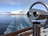 A scenic view of snow-capped mountains across calm ocean waters, as seen from the deck of a cruise ship. A metal binocular stand is prominently displayed in the foreground, suggesting the potential for sightseeing and observation.