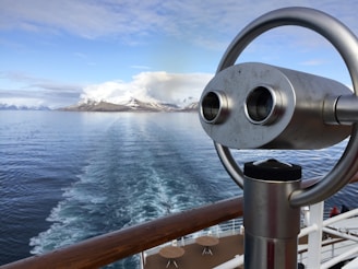 A scenic view of snow-capped mountains across calm ocean waters, as seen from the deck of a cruise ship. A metal binocular stand is prominently displayed in the foreground, suggesting the potential for sightseeing and observation.
