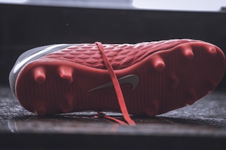 A close-up view of the underside of a red soccer cleat, resting on a reflective surface. The cleat features several studs for grip and a visible swoosh logo on the side. The laces are also visible, laid untied on the surface.