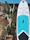 A large paddleboard with a white and blue design is propped upright against a light-colored wall. Next to the paddleboard, there is a tall potted plant with broad green leaves casting shadows on the wall. The background features a clear blue sky with part of a roof visible, suggesting a sunny day.