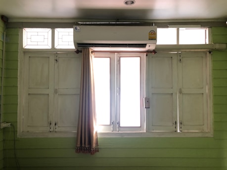 A set of white wooden window shutters with glass panels at the top. An air conditioning unit is mounted above the windows. A beige curtain is partially drawn to the side. The room has pale green walls made of horizontal panels.