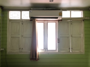 A set of white wooden window shutters with glass panels at the top. An air conditioning unit is mounted above the windows. A beige curtain is partially drawn to the side. The room has pale green walls made of horizontal panels.