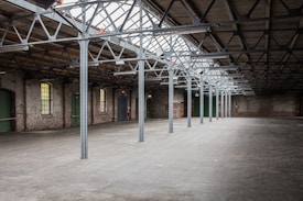 A spacious, empty warehouse with metal trusses and high ceilings made of glass panels. The walls are constructed from exposed brick, featuring several large windows and doors with green accents. The concrete floor is expansive and unoccupied.