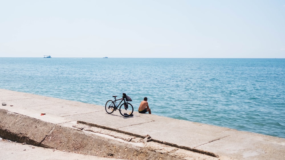 man sitting beside bicycle while sitting on sea wall