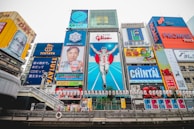 Nighttime cityscape of Osaka illuminated with neon lights, with a tourist capturing the view on their phone.