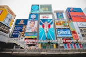 A vibrant cityscape featuring a famous street in Osaka, Japan. The scene is dominated by colorful neon advertisements and billboards, including a large iconic sign of a running man, surrounded by various ads in Japanese. The architecture blends modern and traditional elements, with a river flowing in the foreground and a bridge crossing it.