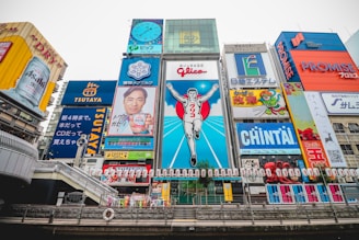 A vibrant cityscape featuring a famous street in Osaka, Japan. The scene is dominated by colorful neon advertisements and billboards, including a large iconic sign of a running man, surrounded by various ads in Japanese. The architecture blends modern and traditional elements, with a river flowing in the foreground and a bridge crossing it.