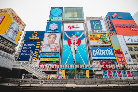 A vibrant cityscape featuring a famous street in Osaka, Japan. The scene is dominated by colorful neon advertisements and billboards, including a large iconic sign of a running man, surrounded by various ads in Japanese. The architecture blends modern and traditional elements, with a river flowing in the foreground and a bridge crossing it.
