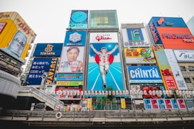 A vibrant cityscape featuring a famous street in Osaka, Japan. The scene is dominated by colorful neon advertisements and billboards, including a large iconic sign of a running man, surrounded by various ads in Japanese. The architecture blends modern and traditional elements, with a river flowing in the foreground and a bridge crossing it.