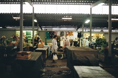 A bustling market with various stalls, including clothing and produce sections. Customers and vendors interact under a roofed structure, with some merchandise displayed prominently.