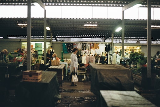 A bustling market with various stalls, including clothing and produce sections. Customers and vendors interact under a roofed structure, with some merchandise displayed prominently.