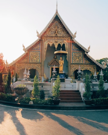 An ornate, golden temple structure with detailed carvings and traditional Thai architecture. A golden statue stands prominently in front, surrounded by various potted plants and decorative elements. The sunlight casts a warm glow on the scene, accentuating the intricate designs and vibrant colors.