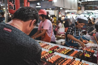 A bustling market scene featuring numerous trays of sushi neatly arranged on a vendor's stall. People are seen selecting sushi, with a vendor actively serving customers. The environment is lively, with other market stalls visible in the background.