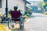 An elderly person with gray hair sits in a red wheelchair on a paved path, surrounded by greenery and buildings in a peaceful outdoor setting.