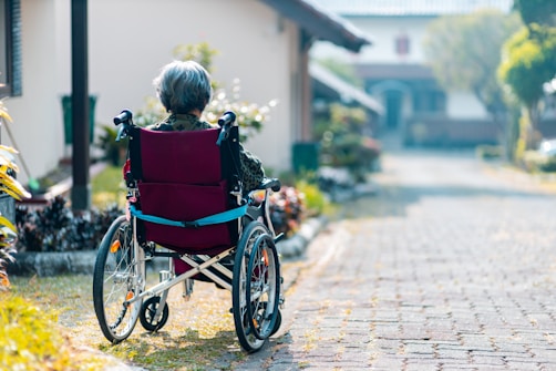 An elderly person with gray hair sits in a red wheelchair on a paved path, surrounded by greenery and buildings in a peaceful outdoor setting.