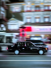A black taxi moves swiftly along a brightly lit urban street at night, with motion blur evident in the background buildings and surrounding vehicles. The illuminated sign on the taxi's roof adds contrast to the otherwise dim evening scene.
