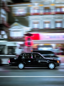 A black taxi moves swiftly along a brightly lit urban street at night, with motion blur evident in the background buildings and surrounding vehicles. The illuminated sign on the taxi's roof adds contrast to the otherwise dim evening scene.
