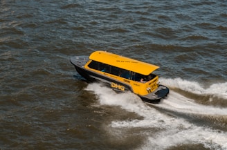 A bright yellow water taxi speeds across choppy water, creating a wake behind it. The taxi is marked with text indicating its service and origin. The scene is dynamic, showing the taxi at an angle with visible movement in the water.