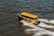 A bright yellow water taxi speeds across choppy water, creating a wake behind it. The taxi is marked with text indicating its service and origin. The scene is dynamic, showing the taxi at an angle with visible movement in the water.