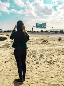 A person stands on a sandy area by the side of a highway, looking towards road signs with multiple lanes visible. The sky is partly cloudy, and the landscape appears dry with sparse vegetation.