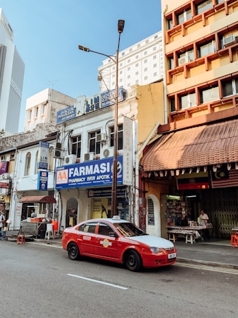 A bustling urban street scene with a row of shops and businesses. A red taxi is parked on the street, while people are seen engaging in various activities outside the shops. The buildings have a mix of architectural styles and display signs in different languages. The sky is clear and blue, and a streetlight stands tall next to the shops.