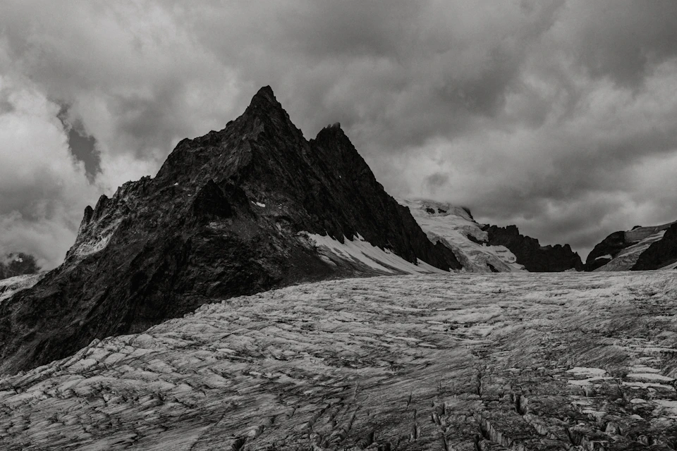 The image depicts a majestic mountain range with two prominent peaks, one larger and more rugged than the other. The mountains are covered in snow and ice, creating a stark contrast against the overcast sky above. The perspective is from below, looking up at the towering heights of the mountains. The black and white color scheme adds a timeless quality to the scene, emphasizing the grandeur and beauty of nature's creations.

The image does not contain any discernible text or human-made objects, making it appear as if we are witnessing these majestic peaks from afar.