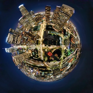 A spherical, panoramic view of a vibrant cityscape at night. Tall skyscrapers with illuminated windows are clustered on one side, creating an urban jungle. Streets with moving vehicles form bright lines and curves, while the city lights create a striking pattern against the dark sky.