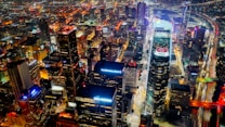 A vibrant nighttime cityscape with illuminated skyscrapers and bustling streets filled with colorful lights. The buildings display various advertisements and logos, adding to the lively urban atmosphere. Streets and highways are visible, showing the movement of traffic as streaks of light.