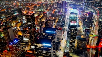 A vibrant nighttime cityscape with illuminated skyscrapers and bustling streets filled with colorful lights. The buildings display various advertisements and logos, adding to the lively urban atmosphere. Streets and highways are visible, showing the movement of traffic as streaks of light.