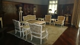 An elegant, classic living room features ornate wooden paneling and vintage furniture. Delicately designed chairs and a sofa, upholstered in patterned fabric, are arranged around a central coffee table atop a large rug. Decorative vases and statues can be seen around the room, along with a grand cabinet against the back wall. Sunlight filters softly through the window curtains, creating a warm and inviting atmosphere.