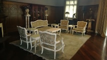 An elegant, classic living room features ornate wooden paneling and vintage furniture. Delicately designed chairs and a sofa, upholstered in patterned fabric, are arranged around a central coffee table atop a large rug. Decorative vases and statues can be seen around the room, along with a grand cabinet against the back wall. Sunlight filters softly through the window curtains, creating a warm and inviting atmosphere.