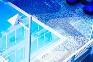 An outdoor swimming pool with clear water reflects the surroundings. The pool area is tiled with small square tiles in varying shades of blue, white, and pink, creating a pixelated effect. There are poolside loungers with dark cushions, and a white metallic structure, possibly for diving or decorative purposes, is prominently visible.