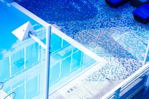 An outdoor swimming pool with clear water reflects the surroundings. The pool area is tiled with small square tiles in varying shades of blue, white, and pink, creating a pixelated effect. There are poolside loungers with dark cushions, and a white metallic structure, possibly for diving or decorative purposes, is prominently visible.
