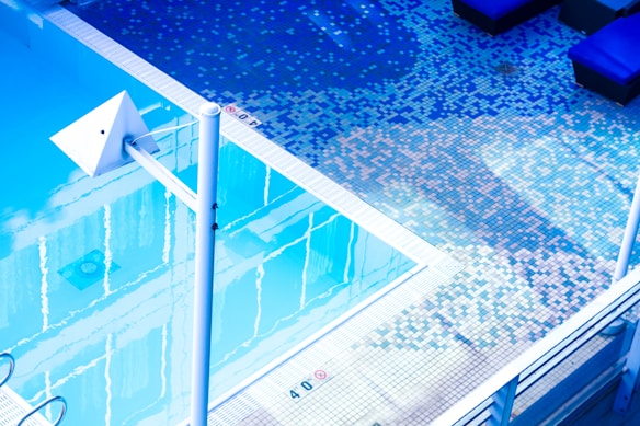 An outdoor swimming pool with clear water reflects the surroundings. The pool area is tiled with small square tiles in varying shades of blue, white, and pink, creating a pixelated effect. There are poolside loungers with dark cushions, and a white metallic structure, possibly for diving or decorative purposes, is prominently visible.