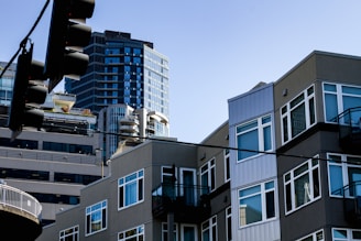 A modern urban scene featuring multi-story residential and commercial buildings with numerous windows. The architectural style is contemporary, with clean lines and a combination of different textures and materials. Tall buildings rise in the background, creating a dense and bustling cityscape. A traffic light hangs in the foreground, partially silhouetted against the clear blue sky.