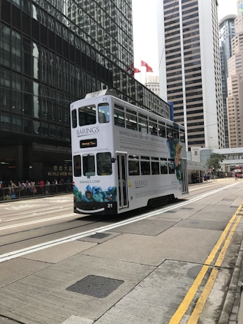 A tram travels along the street in a busy urban setting with tall skyscrapers surrounding it. The tram features an advertisement for Barings, with colorful artwork displayed on its side. The scene includes modern glass buildings and a bustling city atmosphere.