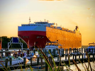 A large cargo ship is docked near a harbor with smaller boats and yachts anchored nearby. The sky is a soft orange hue, suggesting sunset or sunrise, casting a warm glow over the scene. A few people can be seen on the smaller boats in the foreground, enjoying the maritime setting.