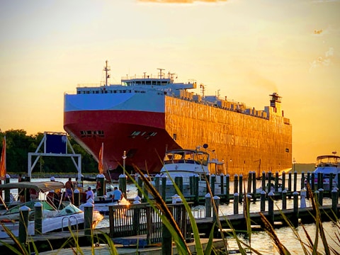 A large cargo ship is docked near a harbor with smaller boats and yachts anchored nearby. The sky is a soft orange hue, suggesting sunset or sunrise, casting a warm glow over the scene. A few people can be seen on the smaller boats in the foreground, enjoying the maritime setting.