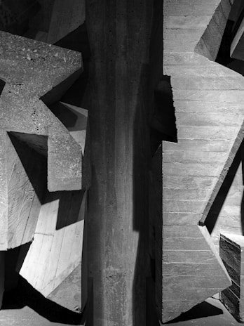 A detailed view of a concrete structure with angular, geometric shapes. The texture of the concrete is rough and varies in tone, creating a play of light and shadow across the surfaces.