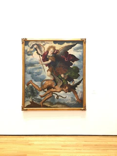 A framed painting depicts a dynamic scene, featuring an angelic figure with wings and a sword confronting a demonic figure. The angel is portrayed flying through a cloudy sky, wearing a garment with red and blue hues. The demonic figure appears in a submissive position below. The painting is set in an ornate gold frame, displayed against a plain white wall with a wooden floor beneath.