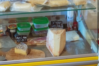 A refrigerated display case filled with various types of cheese, some packaged and some displayed on wooden boards. Visible price labels indicate specific cheeses, alongside containers of deli items. The case also contains plastic containers with green lids and other deli products such as meat slices.