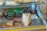 A refrigerated display case filled with various types of cheese, some packaged and some displayed on wooden boards. Visible price labels indicate specific cheeses, alongside containers of deli items. The case also contains plastic containers with green lids and other deli products such as meat slices.