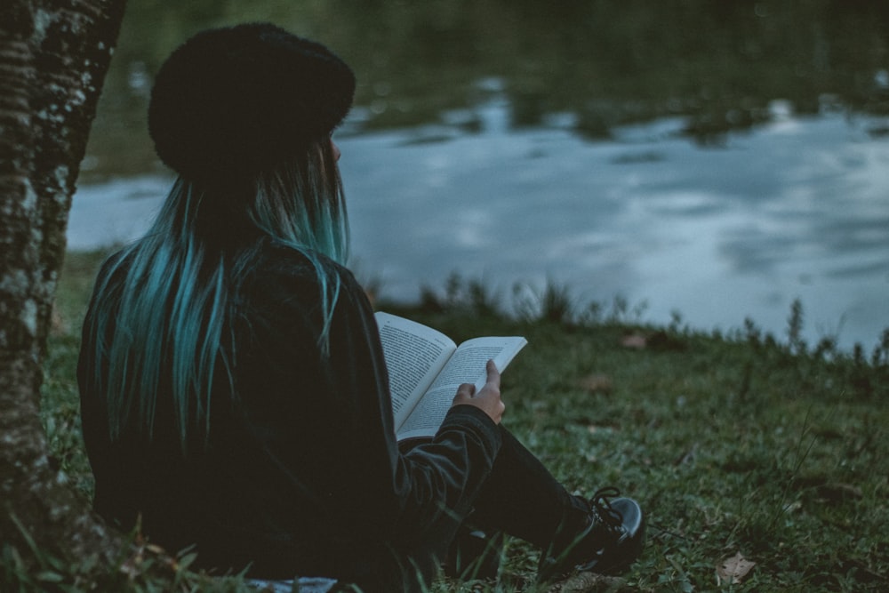 woman holding book while sitting near green field beside lake