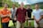 Five men are standing outdoors in front of a wooden fence with a grassy field and forested hills in the background. The men display various casual poses and attire, suggesting a relaxed and friendly atmosphere.