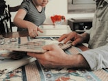 People are sitting at a table engaging in an activity involving photo albums. A person in a striped shirt is pointing at something, while another person is holding the album. There are decorative red objects on the table, likely flowers, and a patterned tablecloth.