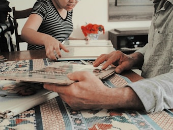 People are sitting at a table engaging in an activity involving photo albums. A person in a striped shirt is pointing at something, while another person is holding the album. There are decorative red objects on the table, likely flowers, and a patterned tablecloth.