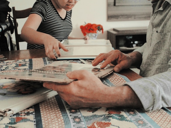 People are sitting at a table engaging in an activity involving photo albums. A person in a striped shirt is pointing at something, while another person is holding the album. There are decorative red objects on the table, likely flowers, and a patterned tablecloth.