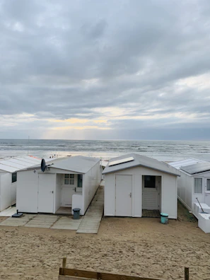 White beach cabins with flat roofs stand in the foreground on a sandy surface with wooden walkways. The cabins are neatly aligned, facing the ocean which is in the background. The ocean waves gently meet the shore. The sky is overcast with dense clouds, allowing only a sliver of sunlight to break through, illuminating the horizon.